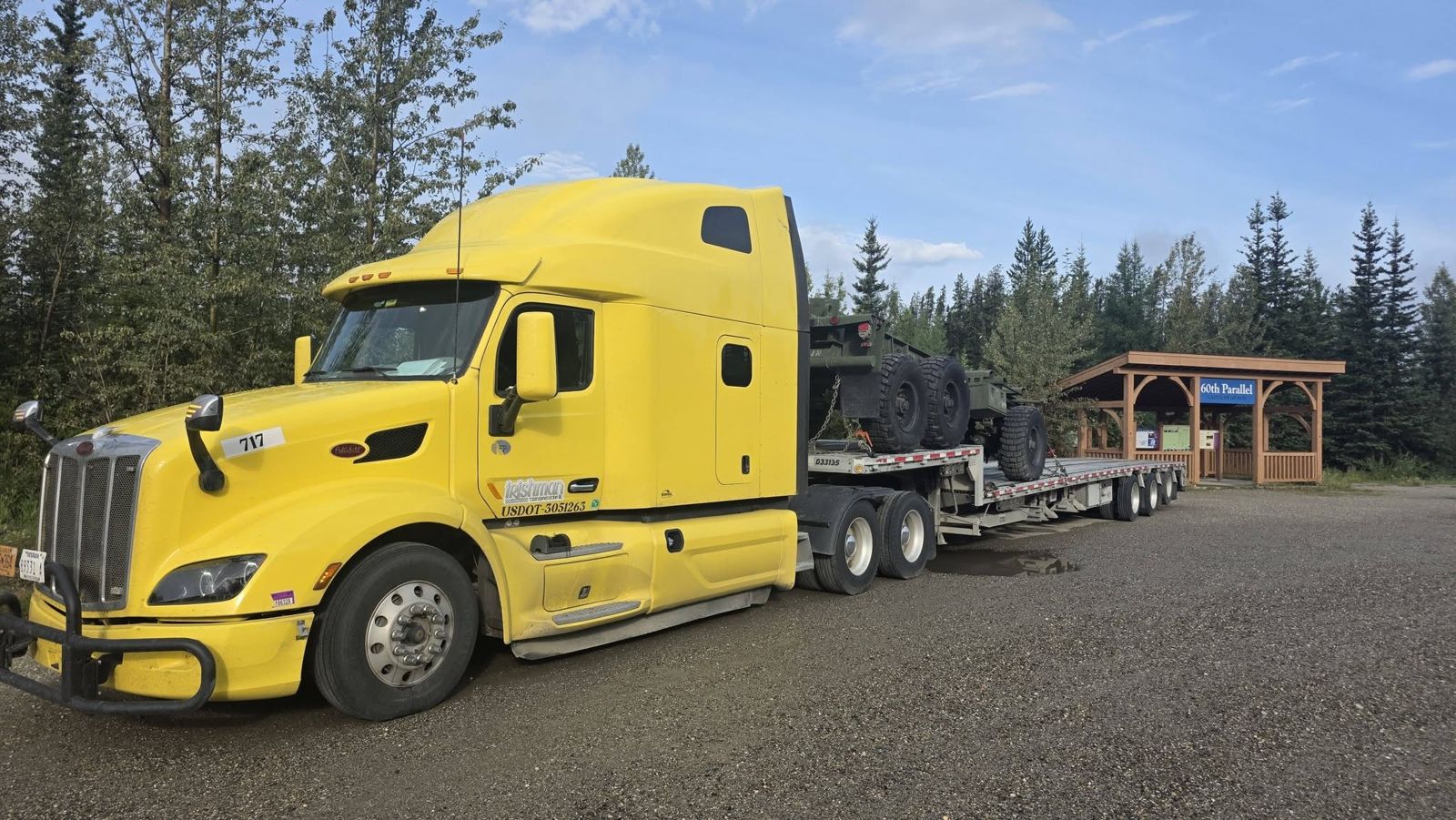Yellow 2020 Peterbilt 579 hauling a military truck at the 60th Parallel sign, Watson Lake, Yukon